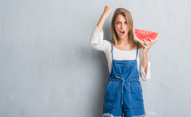 Beautiful young woman over grunge grey wall eating water melon annoyed and frustrated shouting with anger, crazy and yelling with raised hand, anger concept