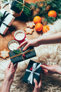 partial view of friends exchanging wrapped christmas gifts at backdrop with cups of cococa drinks and tangerines