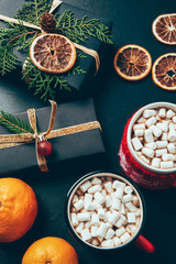 top view of cups of hot drinks with marshmallows, tangerines and christmas presents on black surface