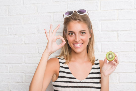 Beautiful young woman over white brick wall eating green kiwi doing ok sign with fingers, excellent symbol