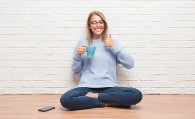 Beautiful young woman sitting on the floor driking cup of coffee at home happy with big smile doing ok sign, thumb up with fingers, excellent sign