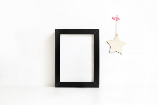 Vertical black blank wooden frame mockup on white table. Wooden star decoration hanging on the wall. Styled stock feminine photography. Home decor. Christmas winter concept.