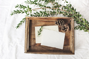 Christmas or winter wedding mock-up scene. Blank cotton paper greeting cards, old wooden tray, pine cones and green Eucalyptus parvifolia branch.White bed linen background. Flat lay, top view.