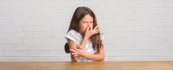 Young hispanic kid sitting on the table at home smelling something stinky and disgusting, intolerable smell, holding breath with fingers on nose. Bad smells concept. - obrazy, fototapety, plakaty