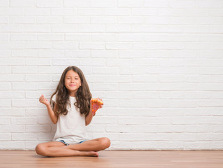 Young hispanic kid sitting on the floor over white brick wall eating pizza slice screaming proud and celebrating victory and success very excited, cheering emotion