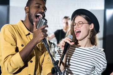 Waist up portrait of two young people enjoying singing with microphones during band performance on...