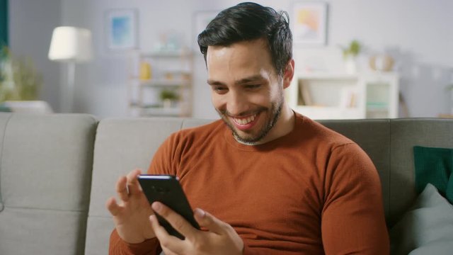 Happy Young Man Uses Smartphone while Sitting on a Sofa at Home