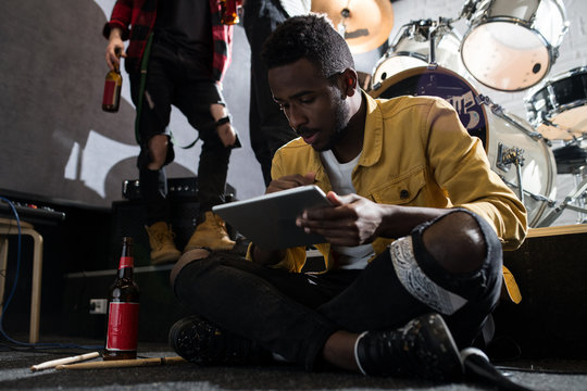 Full length portrait of contemporary African-American man sitting on floor using digital tablet during band rehearsal in studio, copy space