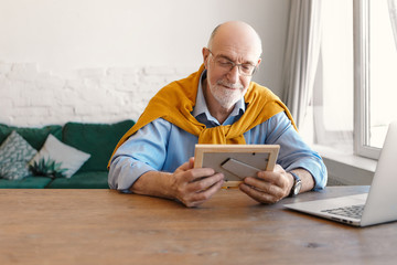 Indoor image of sentimental bearded bald grandfather wearing spectacles and elegant clothes sitting at wooden desk in front of open laptop, holding photo frame, looking at picture of his children