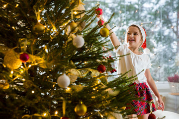 Cute little girl in red Santa hat and plaid skirt decorating Christmas tree at home. Cozy living room decorated with lanterns and Christmas lights. Scenic winter view of pine trees in snow in window