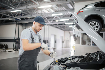 Young worker stands at opened body of car. He uses wrech to fix. He is serious and concentrated. Guy works in garage.