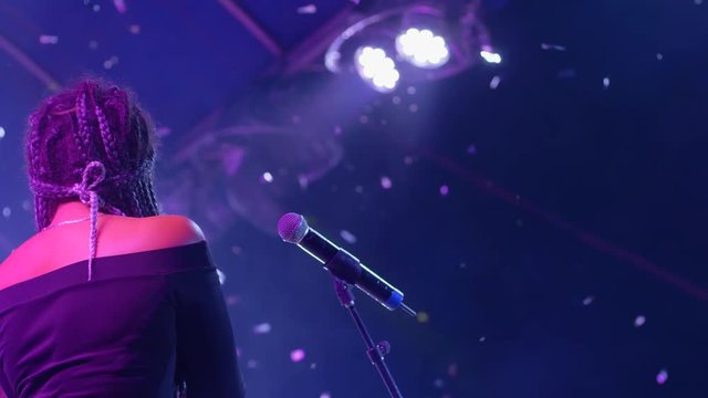 singer woman with afro braces and tambourine into hand sings into microphone on concert stage in bright spotlights