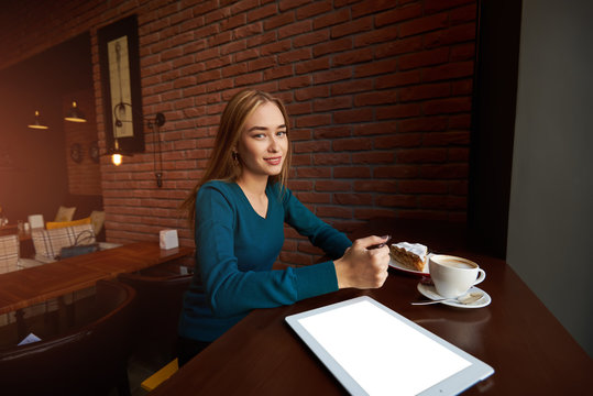 Smiling woman is sitting in comfortable cafe-bar with portable touch pad during her recreation time, cheerful happy female rests after work on digital tablet and waits her friends in cozy coffee shop