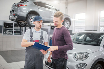Picture of two young men stand in garage together. Bearded guy smiles and points on plastic tablet with documents. Man in grey uniform and cap talk and look at customer. He is serious.