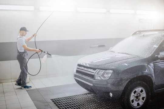 Picture of worker cleaning car. He uses water spray fo that. Young man holds flexible hose. He is serious and concentrated.