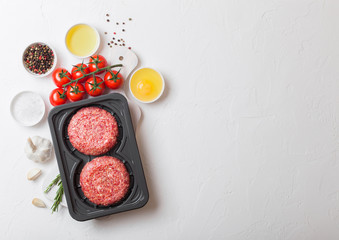 Plastic tray with raw minced homemade beef burgers with spices and herbs. Top view and space for text on top of white kitchen table background with tomatoes salt and pepper.