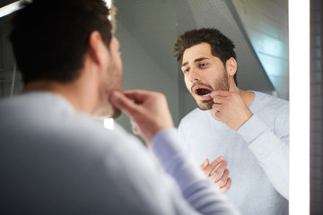 Handsome young man with stubble keeping mouth open while checking tooth and looking into mirror in...