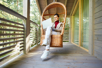 Content confident woman working as freelancer sitting in comfortable hanging chair and using modern...