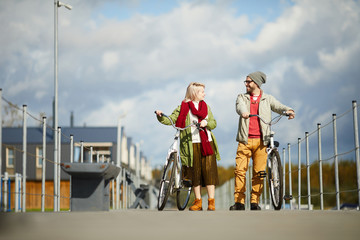 Young stylish Caucasian man and woman walking down the pier with bicycles,  talking and enjoying sunny autumn day, low angle view