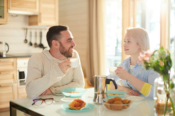 Young married Caucasian couple sitting at table in cozy kitchen, drinking coffee, eating breakfast and talking about plans for weekend