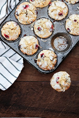 Cranberry Muffins in a muffin tin with kitchen towel over a rustic wood background with scattered fresh cranberries on the table.