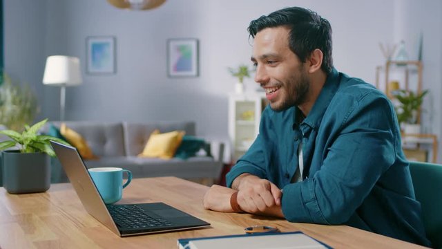 Handsome Young Man Makes Video Call with His Laptop Computer. His Sitting at the Desk in Cozy Living Room.