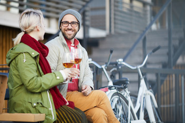 Fototapeta na wymiar Handsome Caucasian bearded man smiling cheerfully while sitting outdoors with his girlfriend and drinking wine on beautiful fall day