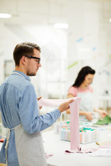 Obraz premium Serious pensive handsome young dressmaker with beard wearing apron and glasses standing at table in workshop and looking at pink fabric while thinking about color for new garment