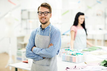 Cheerful confident young male tailor in apron and glasses crossing arms on chest and leaning on table while looking at camera in own atelier