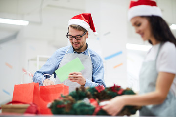 Cheerful positive young man in Santa hat wearing apron and eyeglasses standing at table and putting Christmas card in red paper bag while packaging holiday gifts in workshop