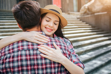 Positive picture of young woman embracing her man. She smiles and keeps eyes closed. She wears hat. They stand on stairs.