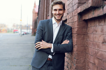 Smiling young male manager formal dressed leaning on a wall outdoors.