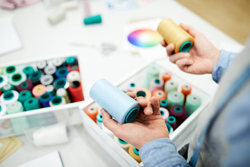 Close-up of unrecognizable male tailor standing at table with containers full of thread spools and thinking of color while choosing thread for sewing