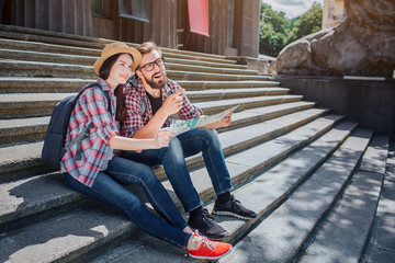 Tourists sit on steps and look forward. Young man points. They smile. People hold map together. It is sunny outside.