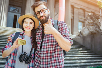 Couple of young tourists stand outisde at stairs and pose on camera. She holds gold card in hand....