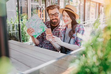 Lovely couple of trvellers sitting and looking at phone woman holds in hands. They smile. Young man holds map in hand. Tourists are surrounded by green plants.