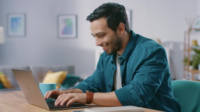 Portrait of Handsome Young Man Using Laptop Computer at Home, Watching and Laughing at Content. Happy Hispanic Man Works on Computer and Drinks from Mug.