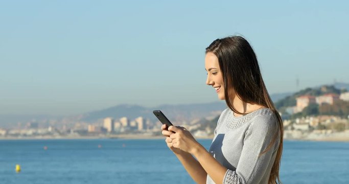 Profile of a happy woman texting on phone standing on the beach