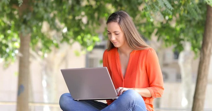 Serious woman uses a laptop writing emails sititng on a bench in a park