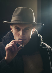 Vintage detective concept. Man in coat, hat smoking cigar, dark background. Macho on mysterious...