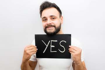 Postitive but strange young man stand and look at camera. He holds black plate with written work yes. Guy holds it with both hands. Isolated on white background.