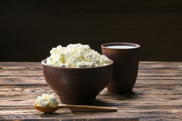 Fresh cottage cheese in  clay bowl with wooden spoon with a glass of milk on rustic wooden background
