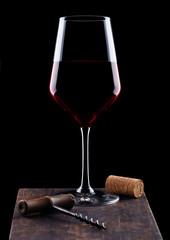 Glass of red wine with vintage corkscrew opener and cork on wooden board on black background.