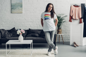 smiling young transgender woman in white t-shirt with pride flag looking at camera at home