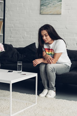 young transgender man sitting on couch at home and looking at pills and water