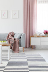 Real photo of bright living room interior with carpet, grey couch with coverlet, window with dirty pink drape and bike-shaped clock on wooden cupboard