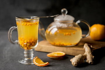 Sea buck thorn tea in glass teapot with glass cup and spoon, orange and ginger near on black background