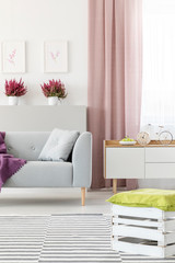 Real photo of white living room interior with couch with pillow, fresh heathers, posters on wall, dirty pink curtain and handmade crate pouf placed on carpet