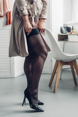 cropped shot of trans man in trench coat putting on stockings at home - obrazy, fototapety, plakaty