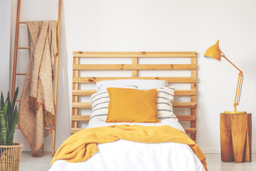 Yellow cushions on wooden bed with blanket in bedroom interior with lamp, plant and ladder. Real...